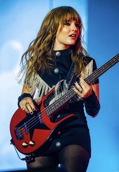 a woman with long hair holding a red bass guitar in front of a blue background