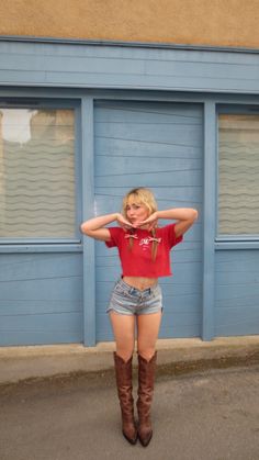 a woman standing in front of a blue building with her hands on her hips and wearing cowboy boots