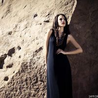 a woman standing in front of a rock wall wearing a black dress and gold necklace