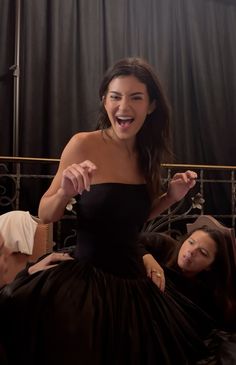 a woman in a black dress laughing while sitting on a bed next to another woman