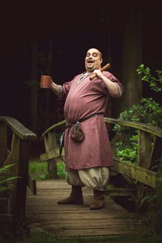 a man dressed in an old fashioned outfit is holding a cup and standing on a wooden bridge