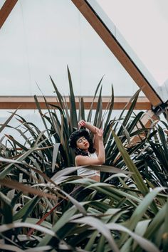 a woman standing in the middle of plants with her hands behind her head and looking at the camera