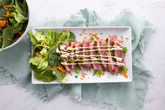 a white plate topped with meat and salad next to a bowl filled with lettuce