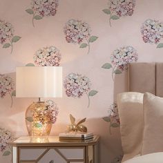 a nightstand with a lamp and flowers on it next to a pink wallpapered bedroom