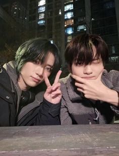 two young men posing for the camera in front of a building at night with their fingers up