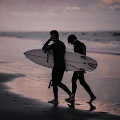 two surfers walking on the beach with their surfboards under their arms as the sun sets