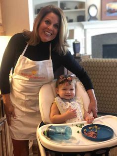 a woman standing next to a baby in a highchair with an apron on