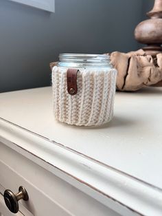 a jar with a knitted lid sitting on top of a dresser next to a candle