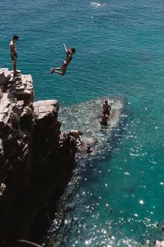 two people jumping off rocks into the ocean from a rocky cliff above them is another person swimming in the water