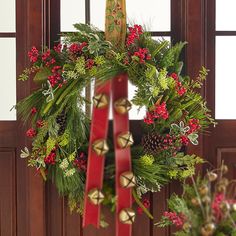 a christmas wreath hanging on the front door with bells and evergreens in red ribbon