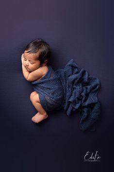 a newborn baby wrapped in a blue blanket is posed on a purple background with her hands behind her head