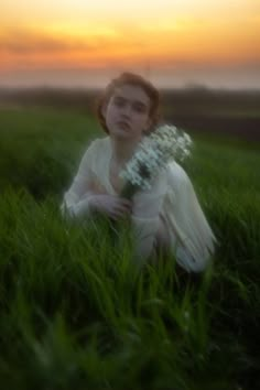 a woman sitting in the grass holding a bouquet of flowers at sunset or dawn with her eyes closed