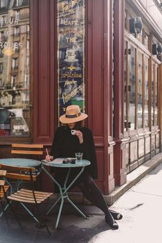 a woman sitting at an outdoor table with a cup of coffee in her hand and wearing a hat