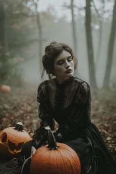 a woman sitting on the ground with two pumpkins in front of her and foggy forest behind her