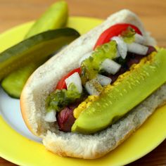 a hot dog with pickles, tomatoes and onions on a yellow plate next to cucumbers