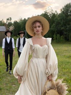 a woman in a white dress and straw hat is holding a basket while two men are walking behind her