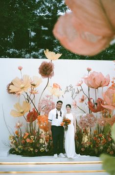 two people standing next to each other in front of flowers and plants on a wall