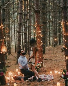 a man kneeling down next to a woman on a blanket in the woods surrounded by candles