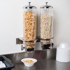 two glass containers filled with cereal on top of a metal counter next to a bowl of cereal