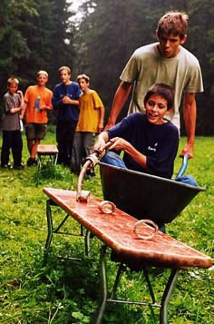 a man pushing a wheelbarrow with two boys in the back and one boy behind him