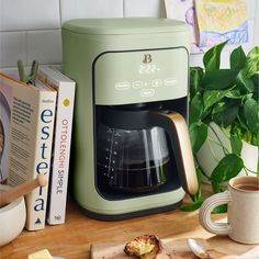 a green coffee maker sitting on top of a wooden cutting board next to a book