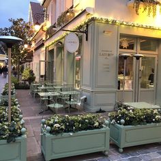 two green planters with flowers in them on the side of a street next to tables and chairs