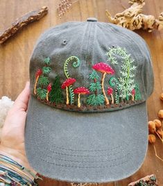 a person holding up a gray hat with embroidered mushrooms and pine trees on the front