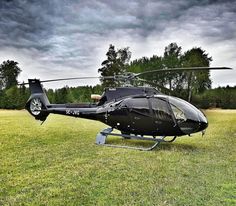 a black helicopter sitting on top of a lush green field next to trees and clouds