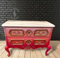 a pink and gold dresser sitting on top of a wooden floor next to a brick wall