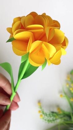 a person is holding a paper flower in front of a plant with yellow flowers on it