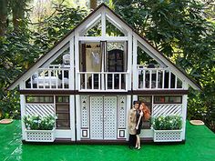 two people standing in front of a doll house on top of a green lawn with trees