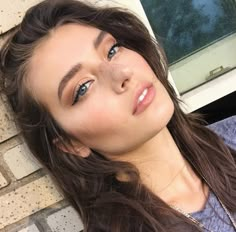 a woman with long brown hair and blue eyes is leaning against a brick wall looking at the camera