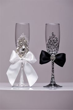 two wine glasses with bows on them sitting side by side, one has a diamond brooch and the other has a black bow