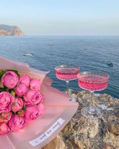 pink roses and two wine glasses sitting on the edge of a cliff overlooking the ocean