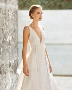 a woman in a white wedding dress standing next to a wall and looking off into the distance