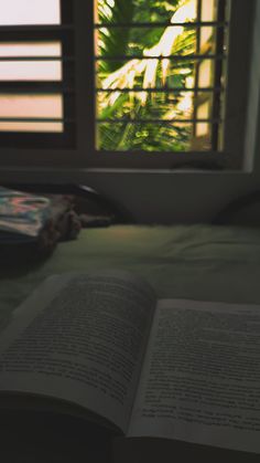 an open book sitting on top of a bed next to a window with shutters