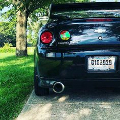 the back end of a black sports car parked in front of a tree and grass area