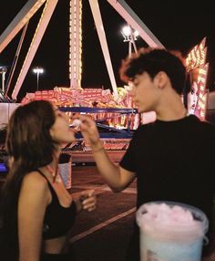 a man and woman standing in front of a carnival ride at night, eating food