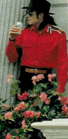 a man in a red shirt and black hat holding a beer next to pink flowers