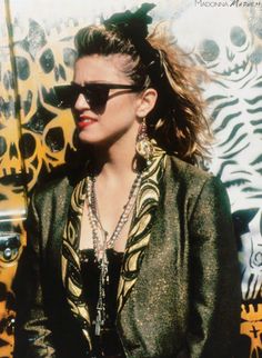 a woman wearing sunglasses and a jacket standing in front of a wall with skulls on it