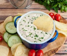 a blue bowl filled with dip surrounded by sliced cucumbers and chips on a wooden board