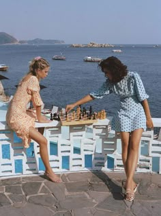 two young women playing chess on a stone patio overlooking the ocean with boats in the background