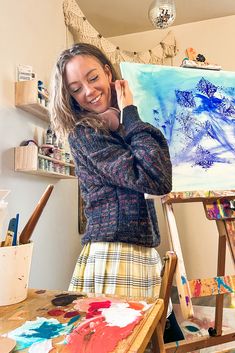 a woman is smiling while painting on an easel in front of her, with one hand up to her face