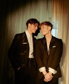 two men standing next to each other in front of a window with curtains behind them