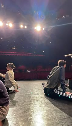 three people sitting on the floor in front of an empty stage with bright lights above them