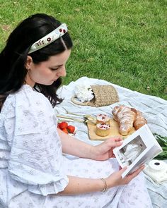 a woman is sitting on the grass reading a book and holding a box with pastries