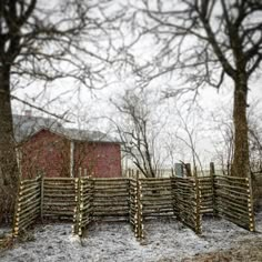 a fence made out of logs in front of a red barn and trees with snow on the ground