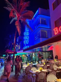 people are walking down the sidewalk in front of a building with neon lights and palm trees