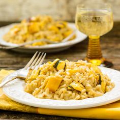 two white plates with rice and squash on them next to a glass of wine in the background