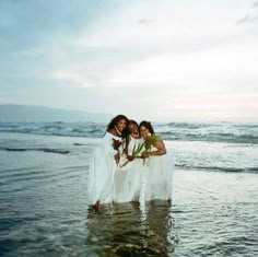 three women in white dresses standing in the water at the beach with their arms around each other